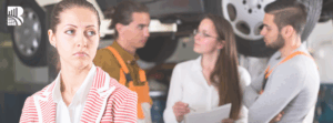 Woman in a striped jacket looking dissatisfied while mechanics and a consultant discuss car service in an auto repair shop – customer service and business excellence concept.