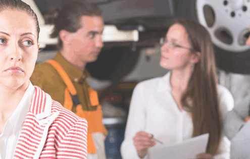 Woman in a striped jacket looking dissatisfied while mechanics and a consultant discuss car service in an auto repair shop – customer service and business excellence concept.
