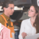 Woman in a striped jacket looking dissatisfied while mechanics and a consultant discuss car service in an auto repair shop – customer service and business excellence concept.