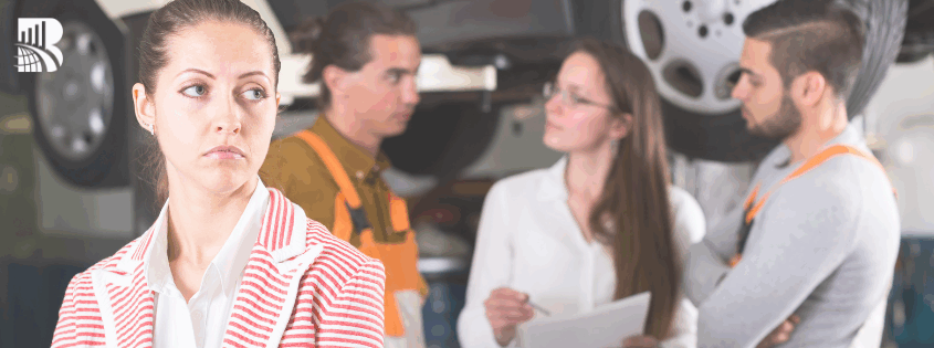 Woman in a striped jacket looking dissatisfied while mechanics and a consultant discuss car service in an auto repair shop – customer service and business excellence concept.